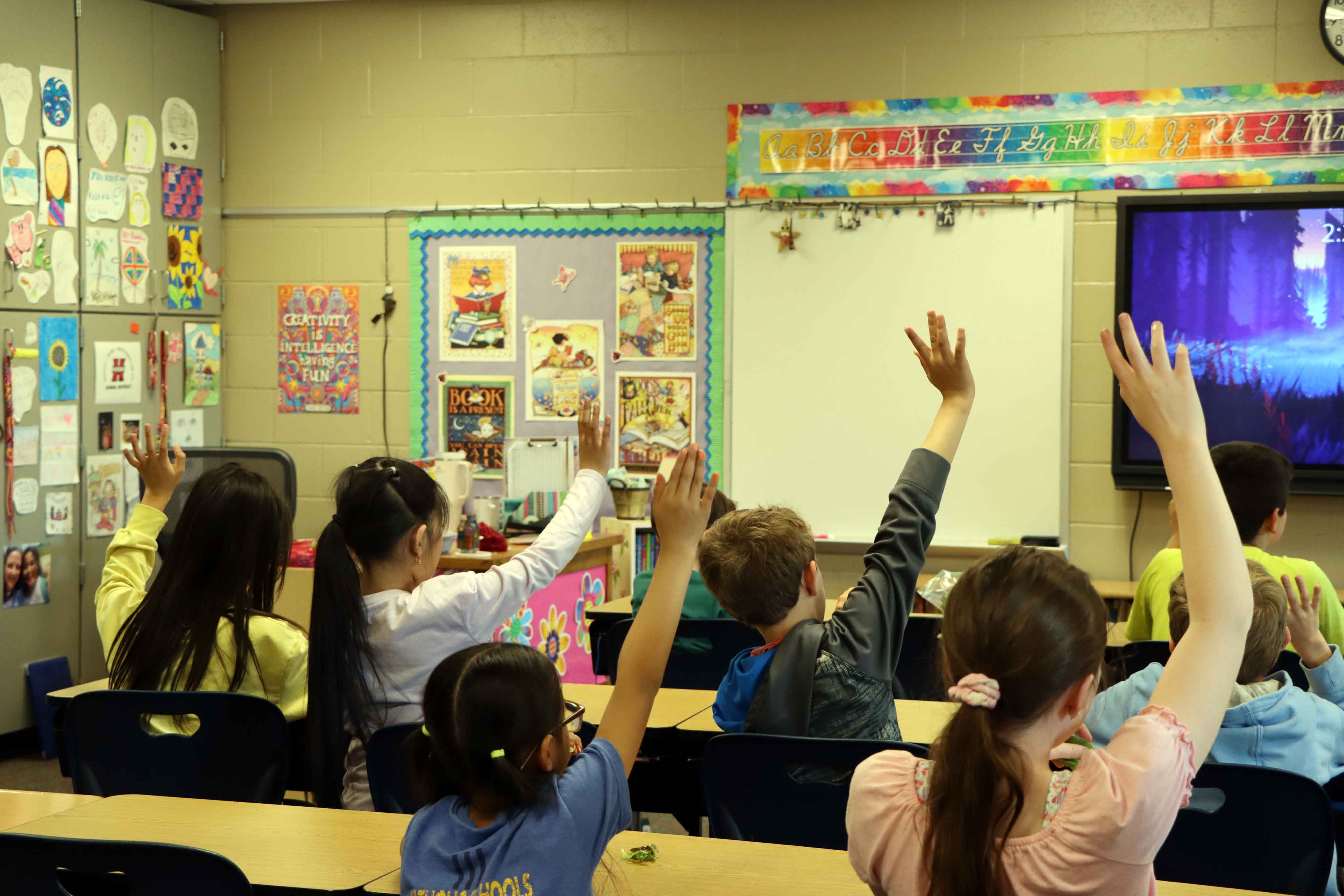 Children seated at desks in a classroom raise their hands