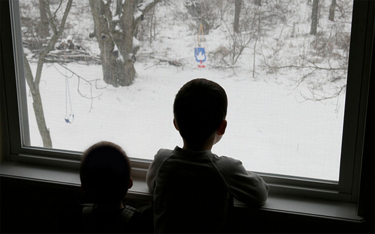 Two children look out their living room window at a snowy winter landscape.