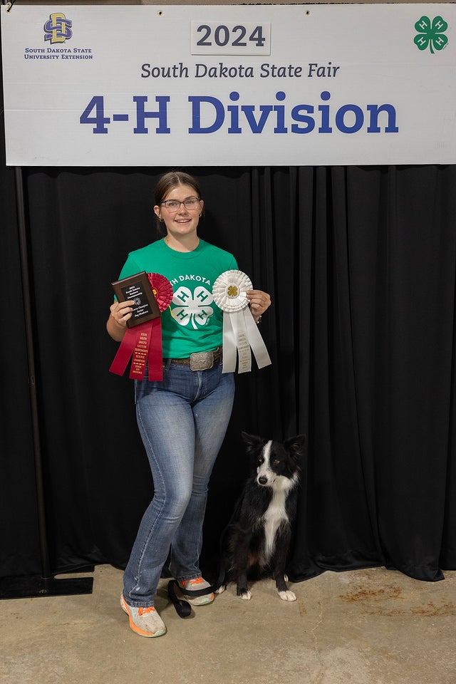 A girl in a green shirt holds up a plaque and large red and white rosettes; a black and white dog sits beside her