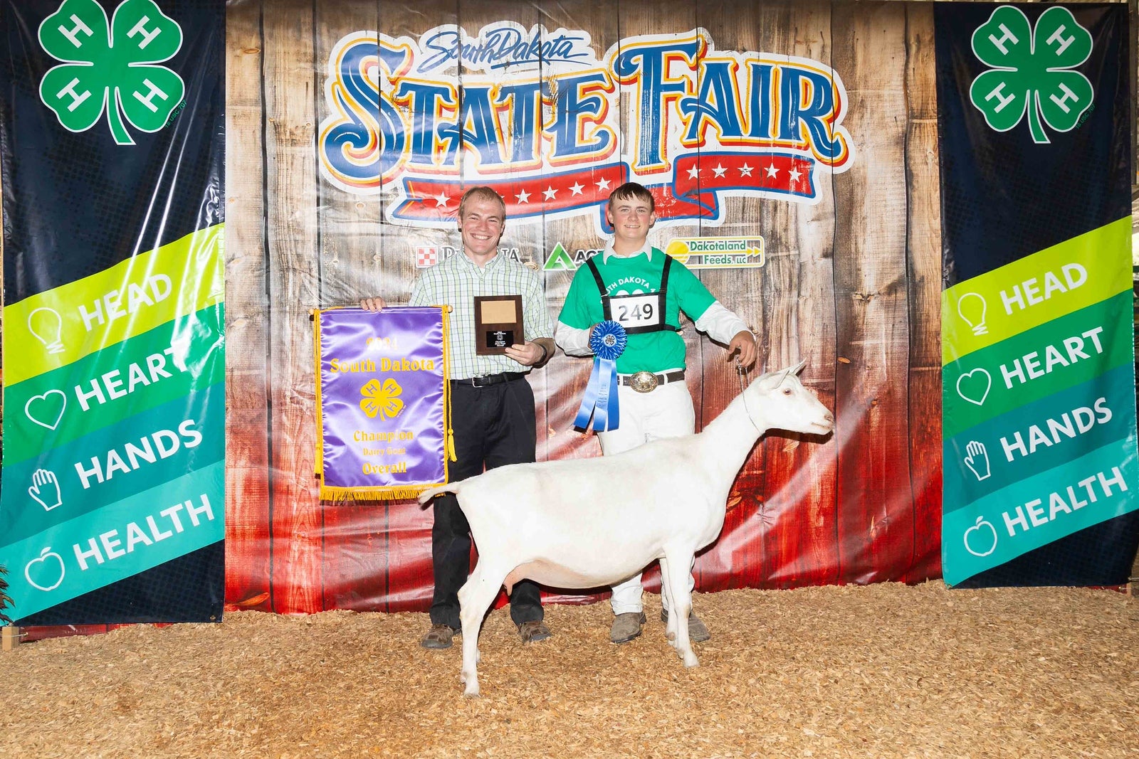A teenage boy holds a goat by the collar and holds up a blue ribbon. Someone beside him is holding up a banner and a plaque