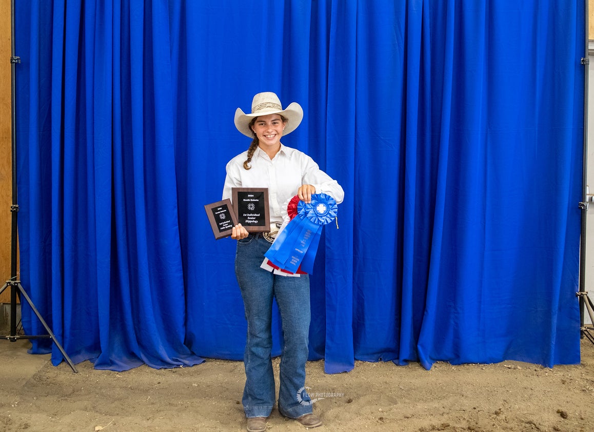 A girl in a cowboy hat holds two plaques and several large rosette awards