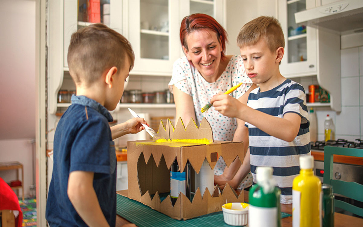 Mother with two sons decorating a dinosaur costume fashioned out of a cardboard box.