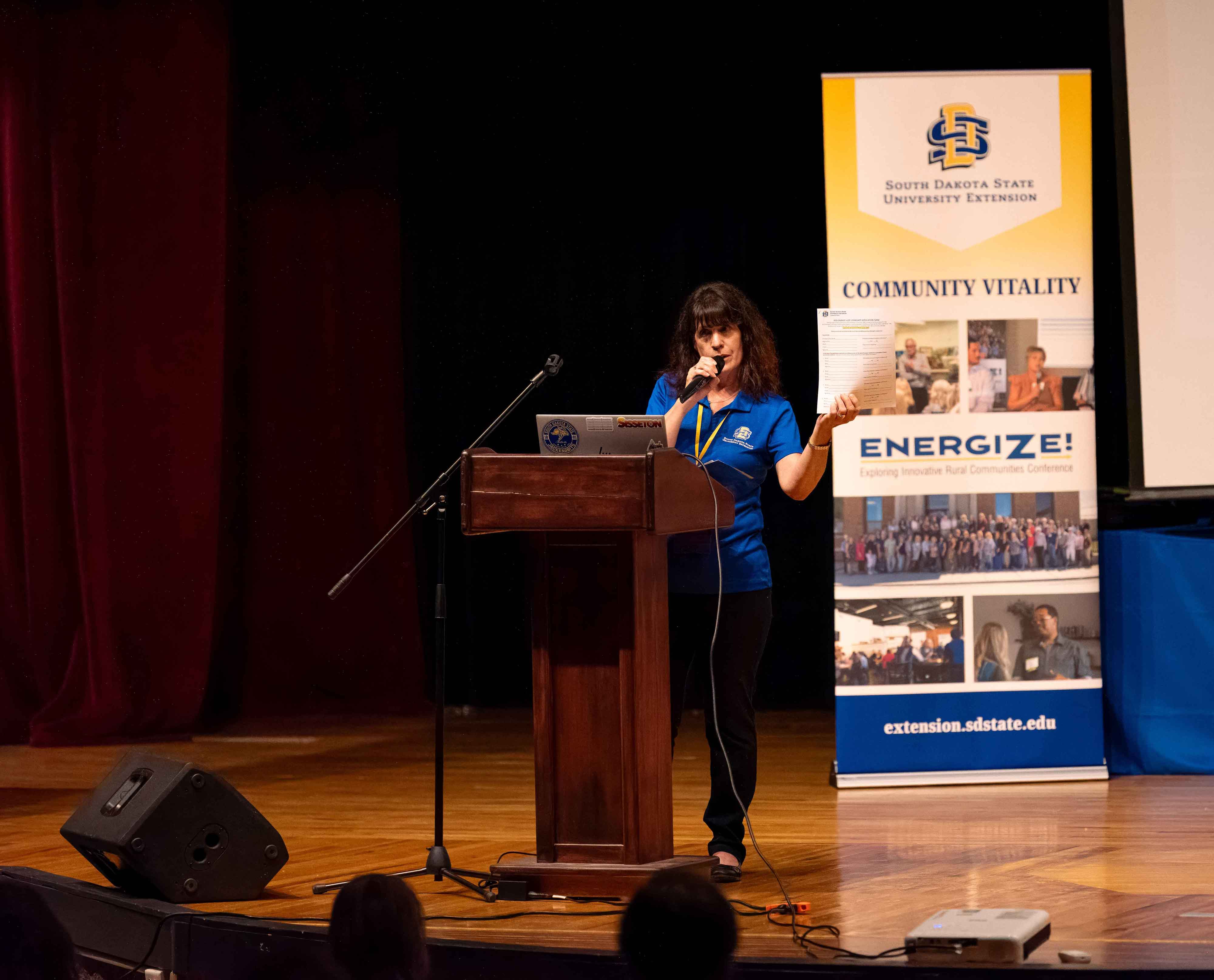 A woman on a stage shows a paper application to her audience, with an Energize banner behind her