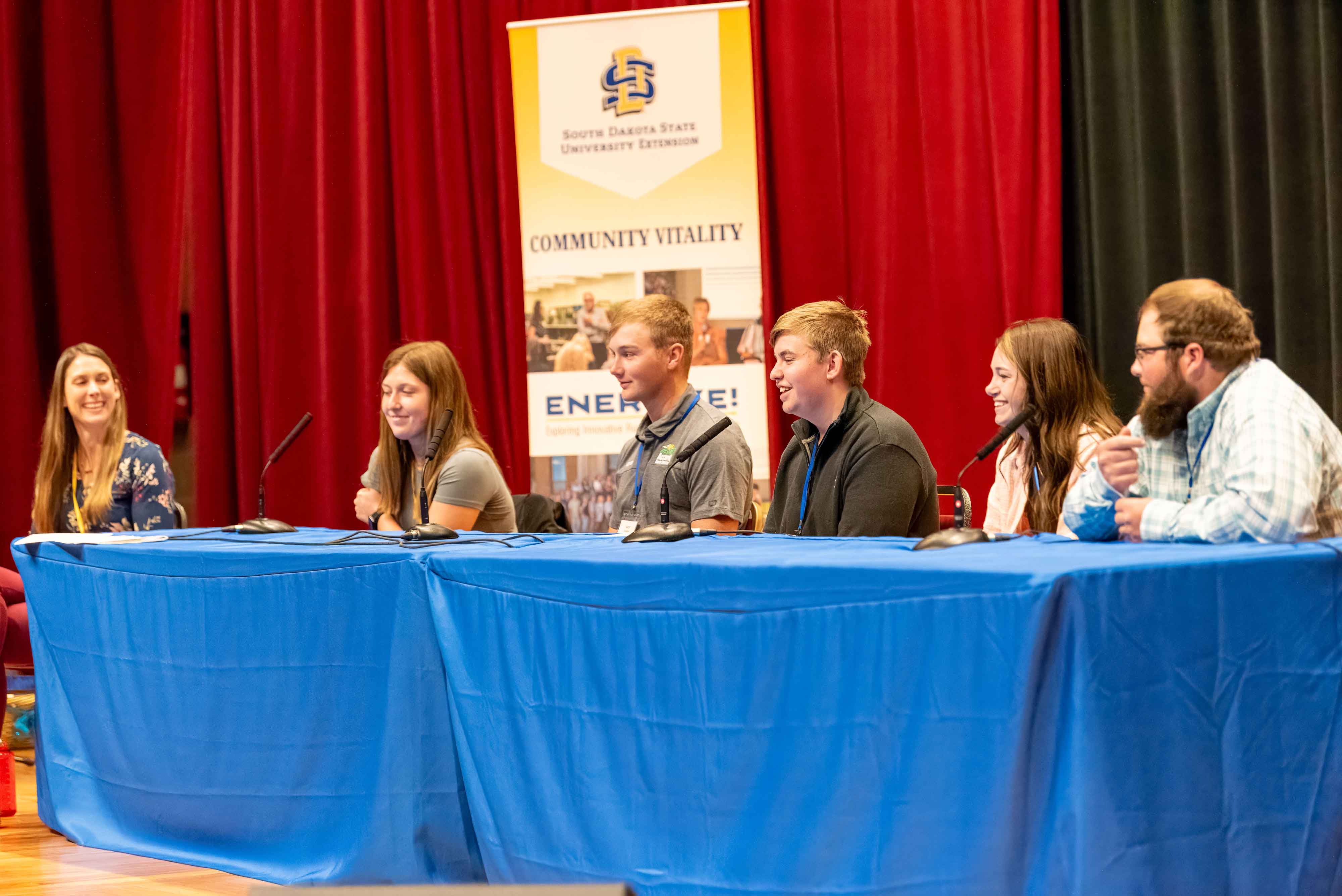A panel of people sits behind tables on a stage with an Energize banner behind them