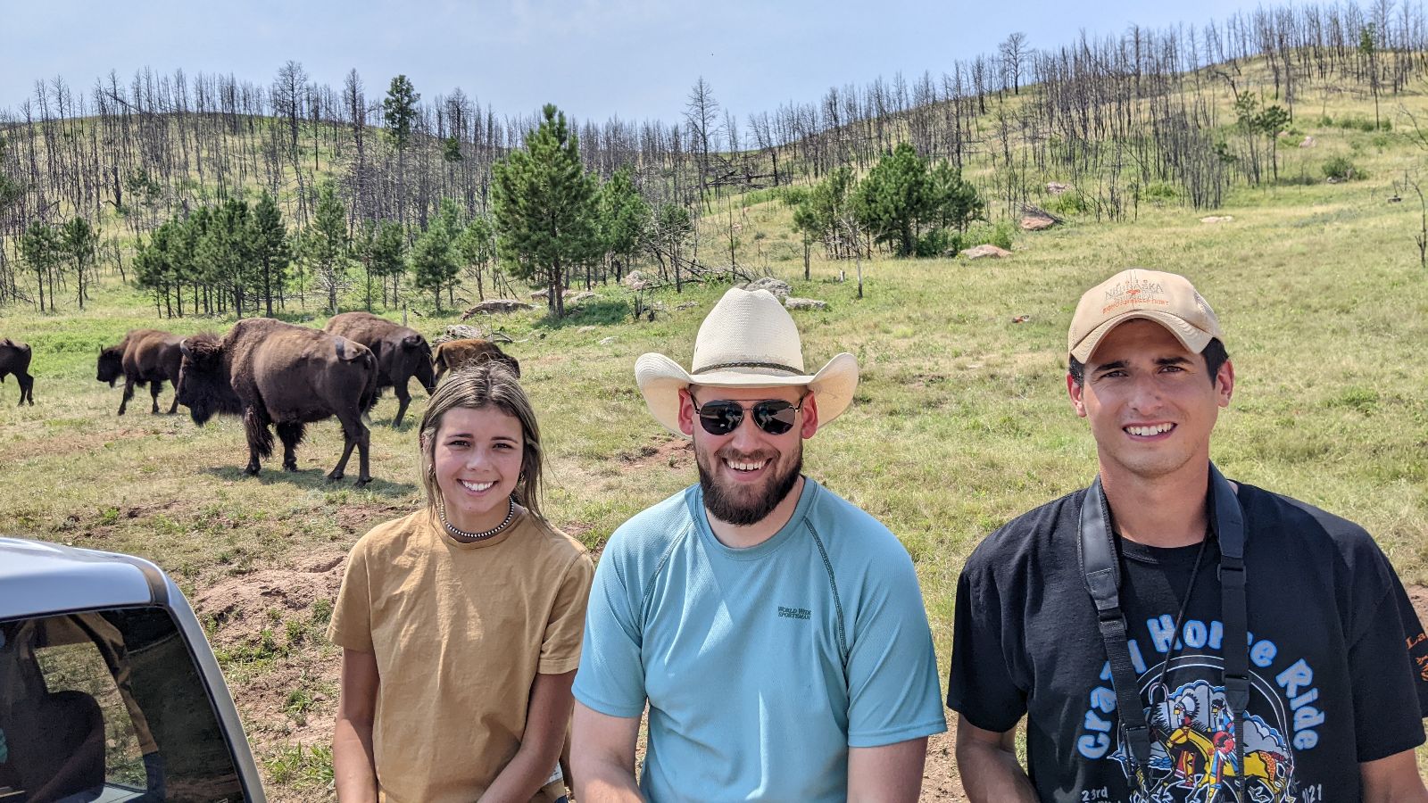 Three people smile for the camera with bison and rolling hills in the background