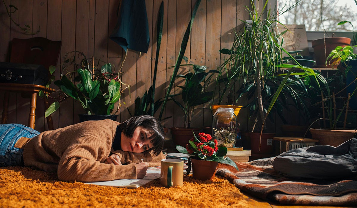 A young woman writing in a notebook with a variety of houseplants in the background.