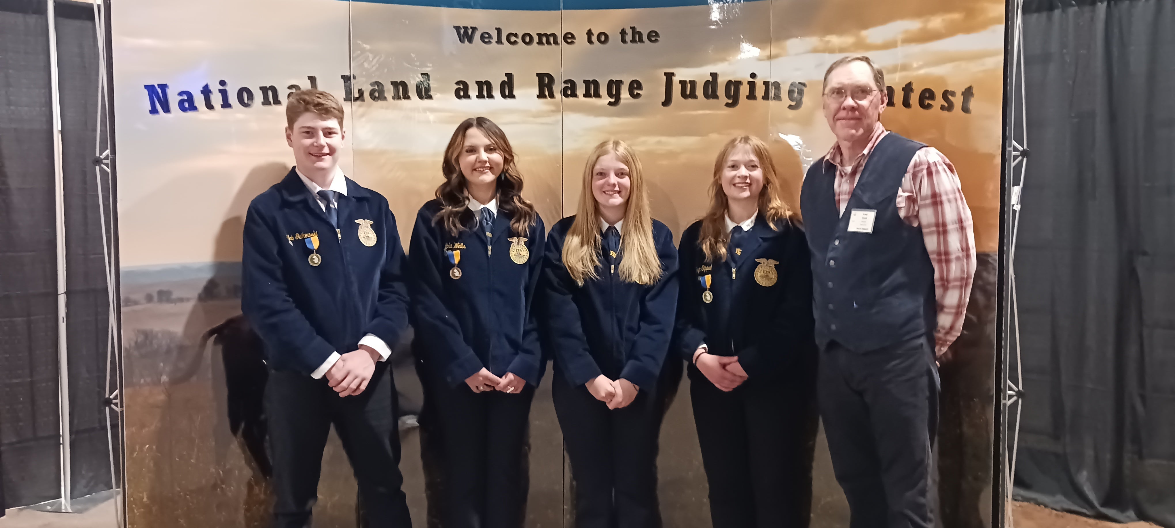 Four youth in blue FFA jackets and their coach pose for a team photo in front of a display that says &quot;Welcome to the National Land and Range Judging Contest&quot;