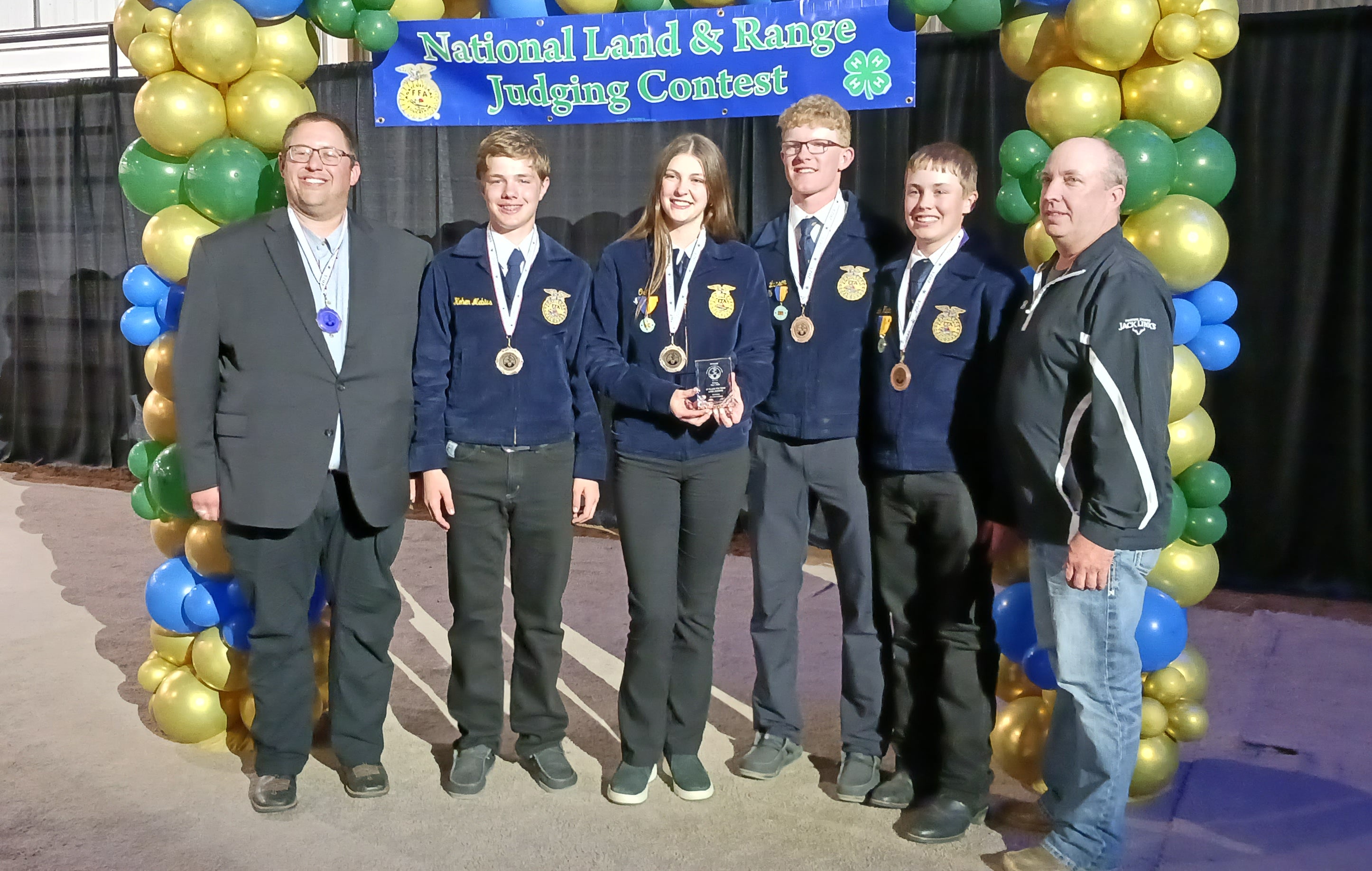 Four youth in blue FFA jackets wear medals around their neck and smile for a team photo with their coaches on either side