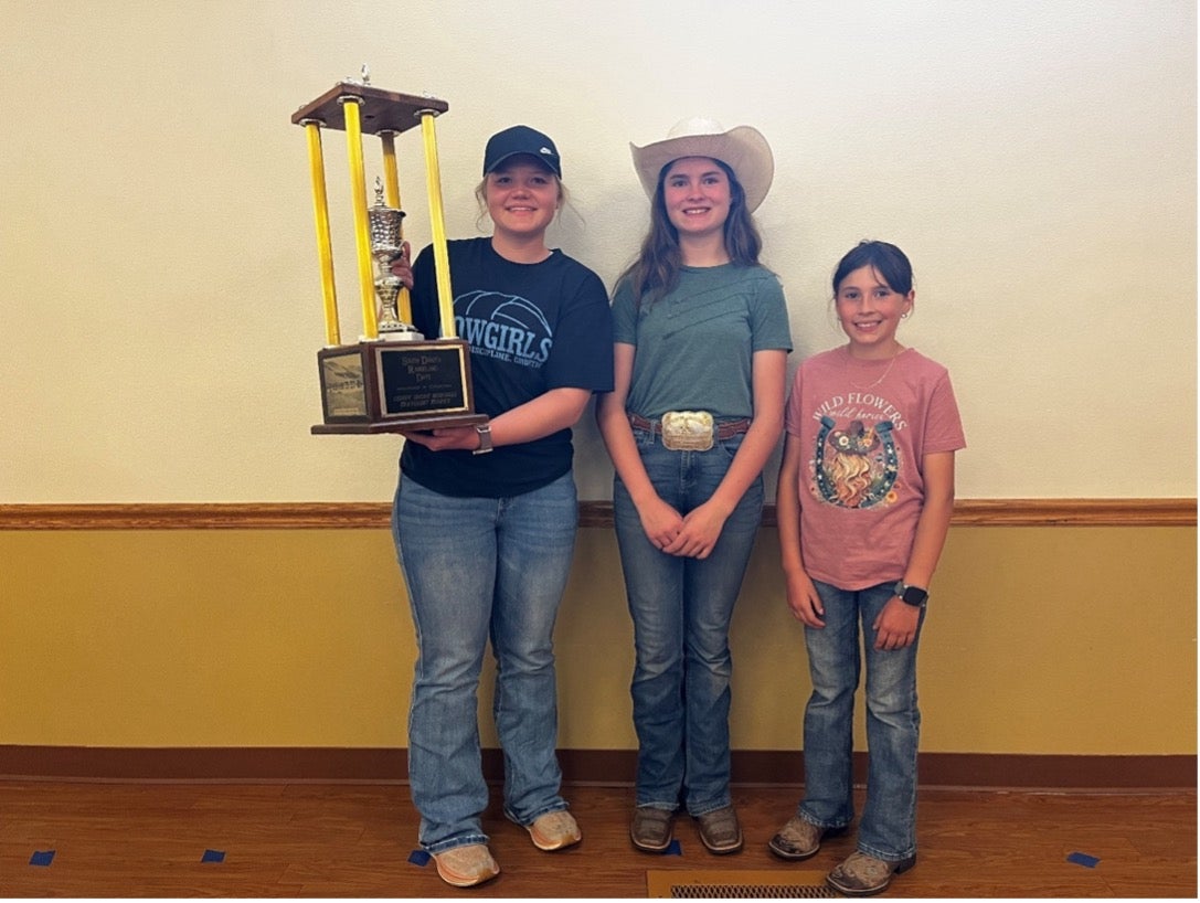 A girl holds a large trophy and two others smile with her for the camera