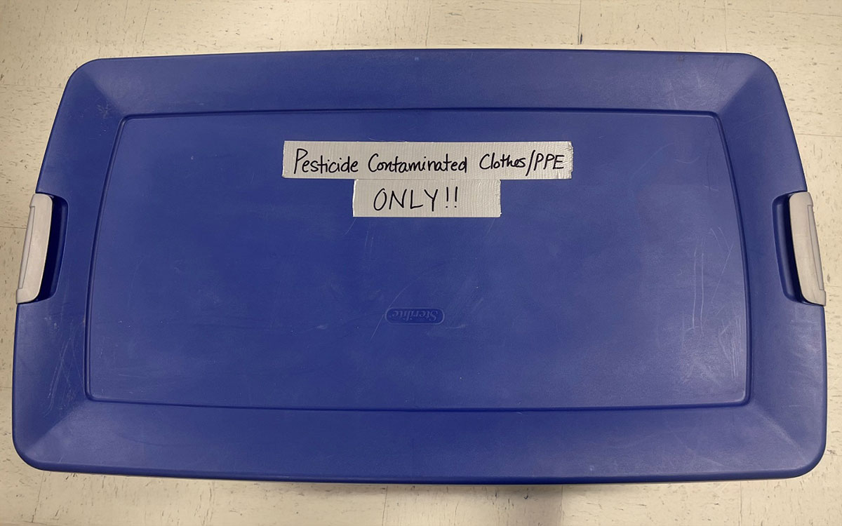 Blue container with lid visible that has a label indicating it is only for pesticide contaminated clothing/PPE.