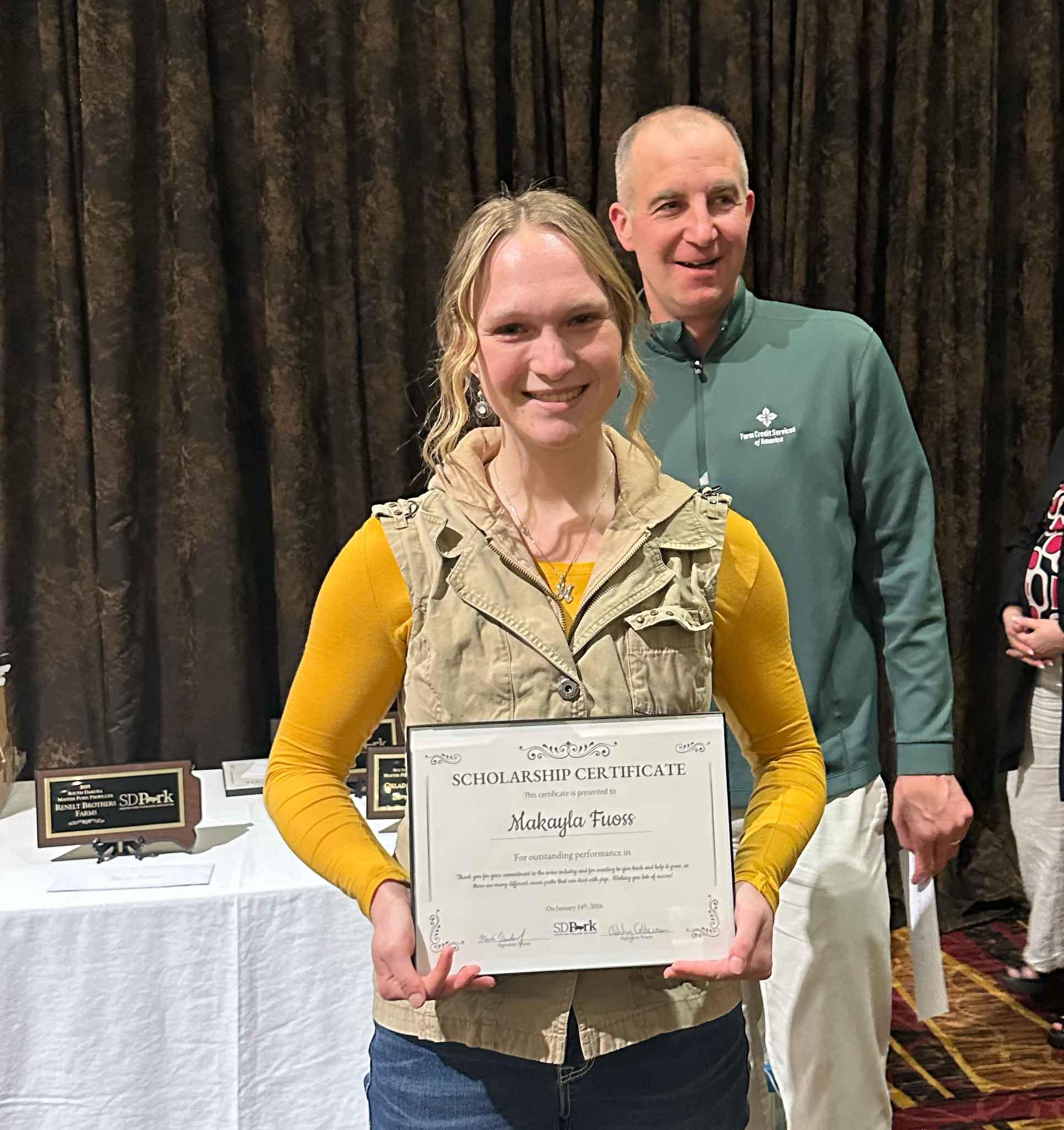 A girl wearing a mustard-colored long-sleeved shirt and beige vest holds up a scholarship certificate with the name "Makayla Fuoss"
