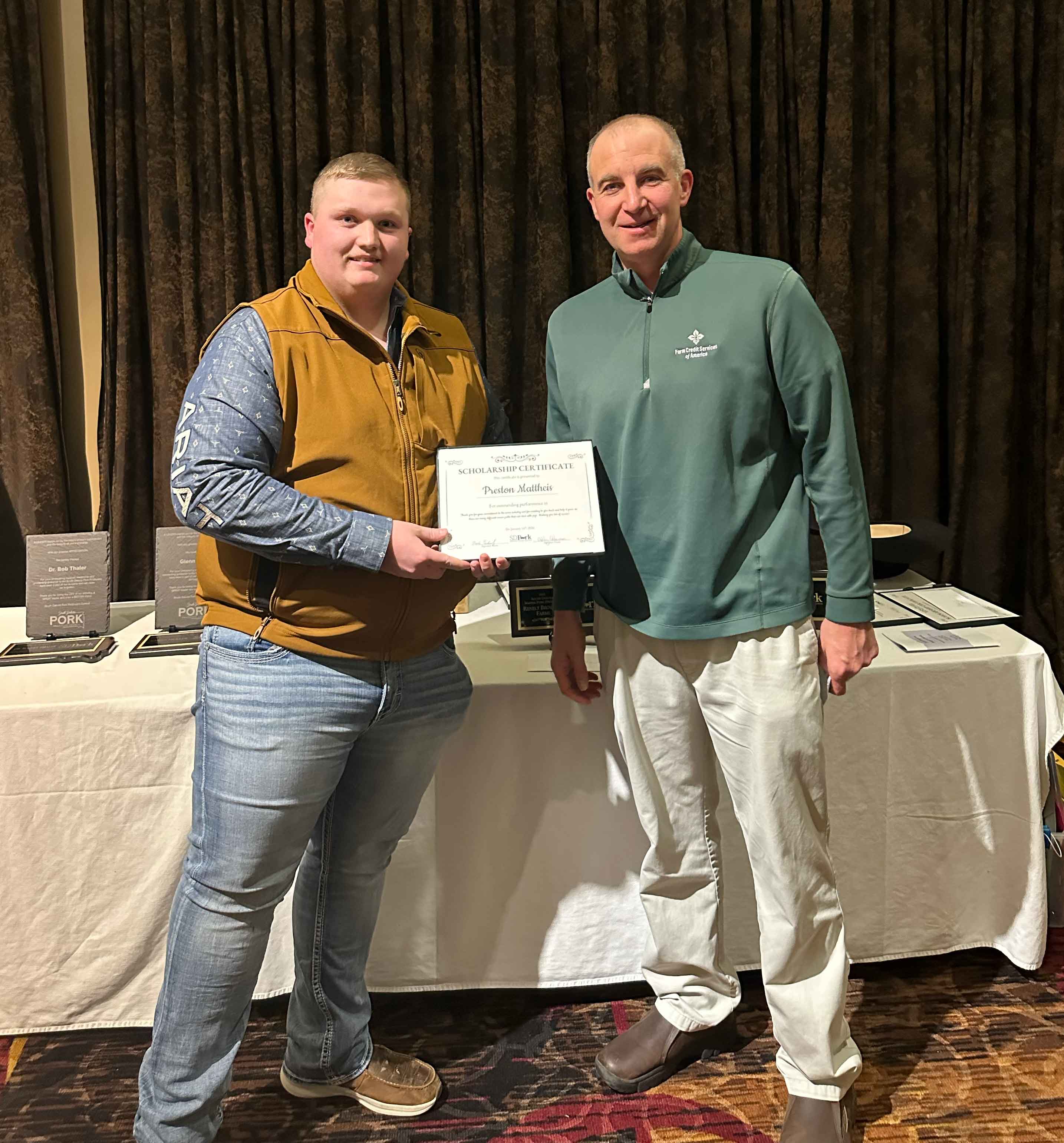 A college student in a long sleeved shirt and orange vest accepts a scholarship certificate from a man in a green shirt