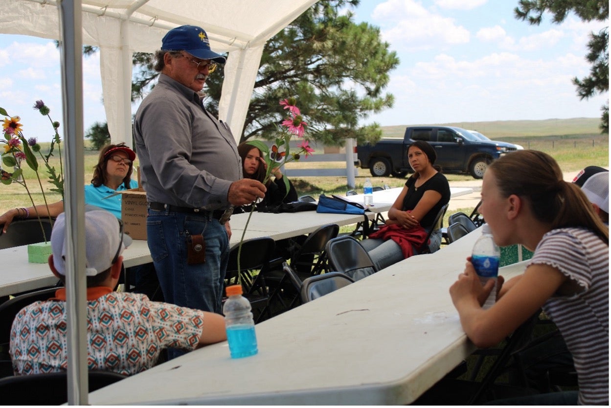 A man holds up a long pink flower stem and talks to a group of youth sitting at long white tables