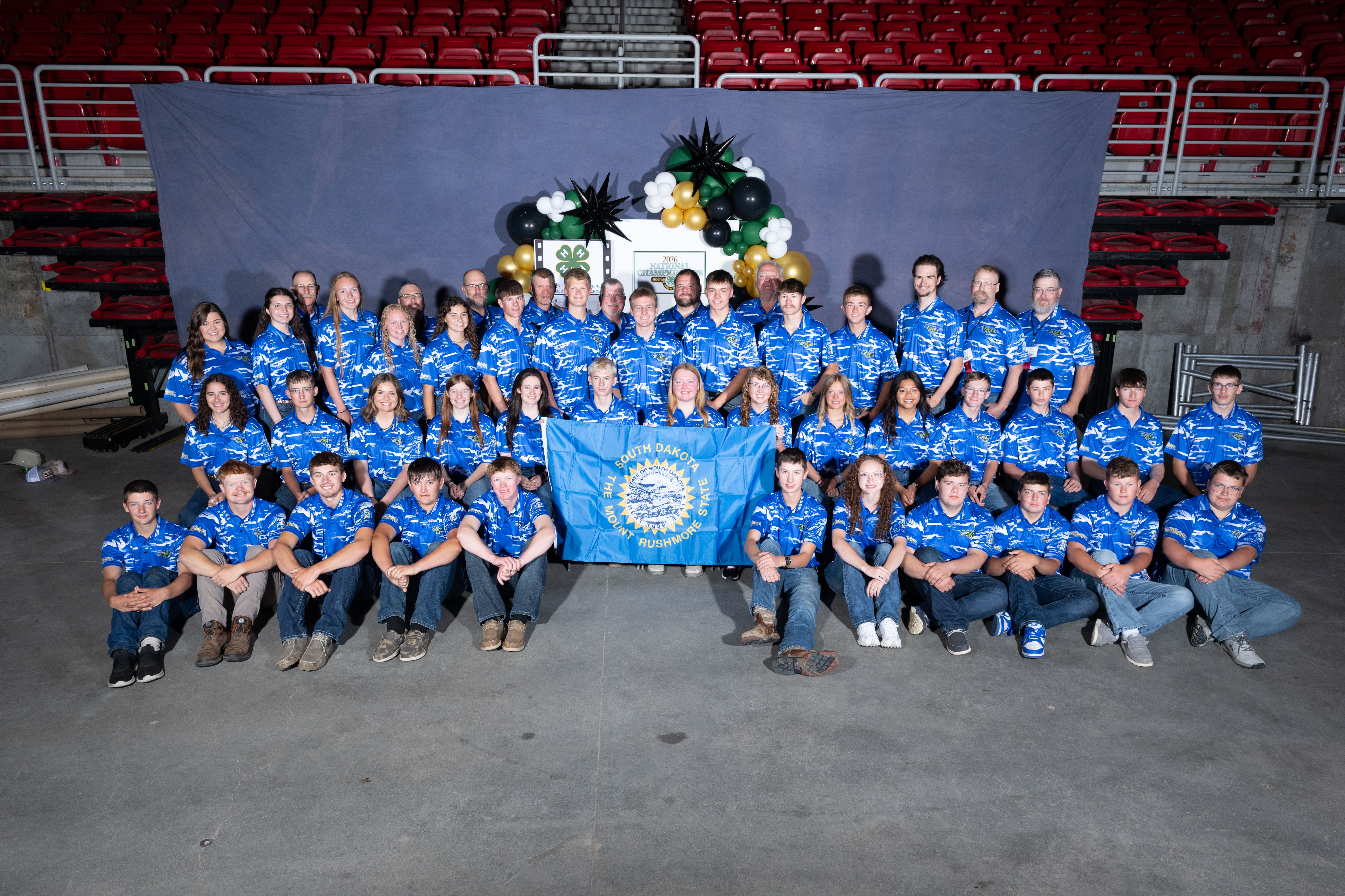 A large group of youth wearing blue T-shirts sits and stands holding a South Dakota state flag