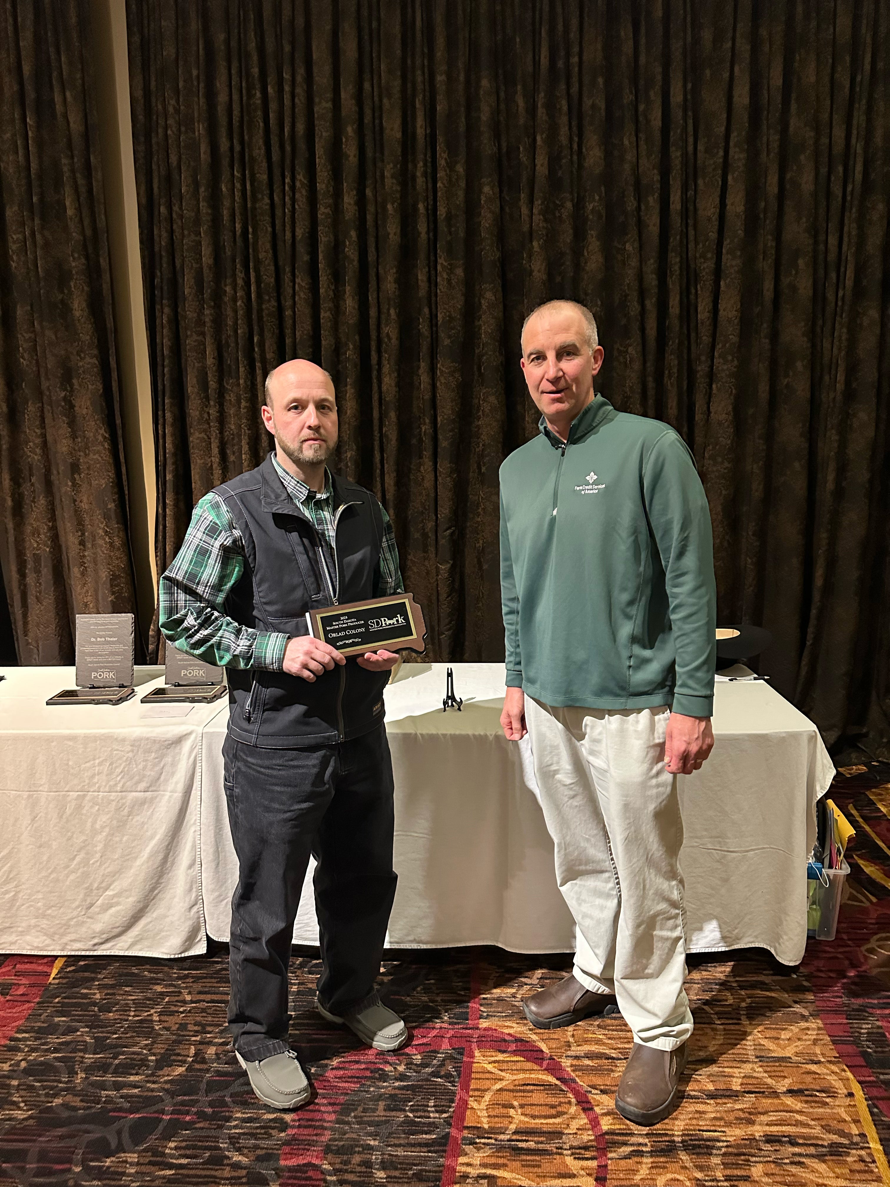 A man in a black vest over a green checkered shirt receives a Master Pork Producer plaque from a tall man in a green long-sleeved shirt