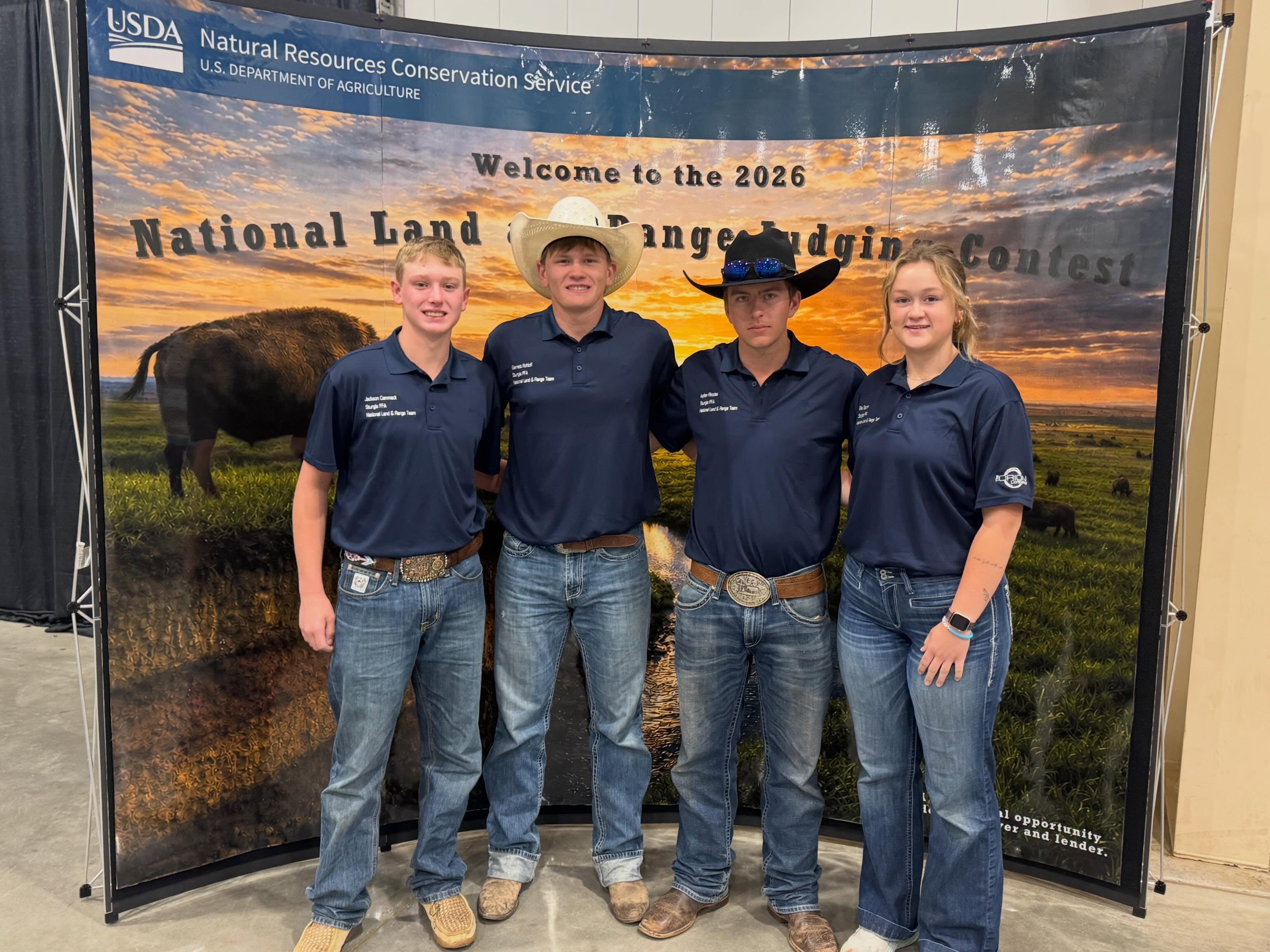 Four people smile in front of a National Land and Range Judging Contest photo banner