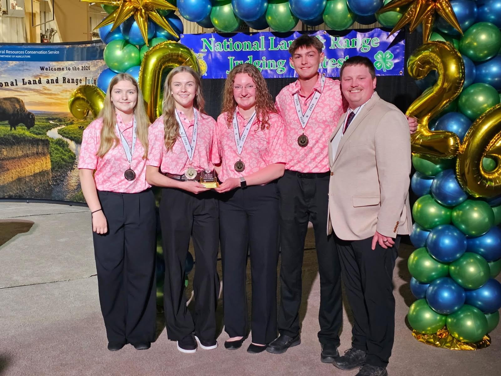 Four peopple in matching pink shirts wear fourth-place medals standing next to their coach in a grey suit jacket