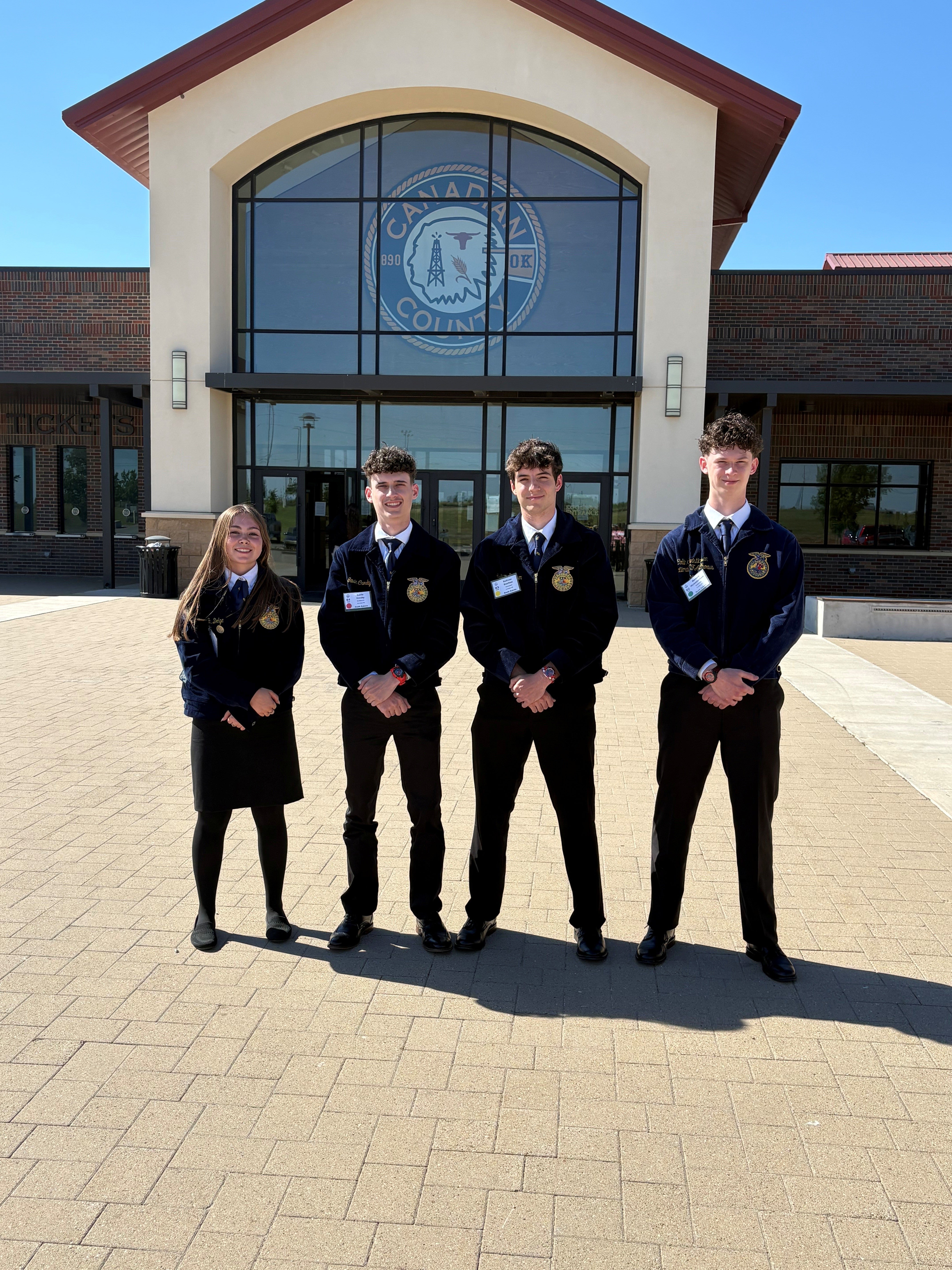 Four youth wearing matching FFA jackets stand outside a large building