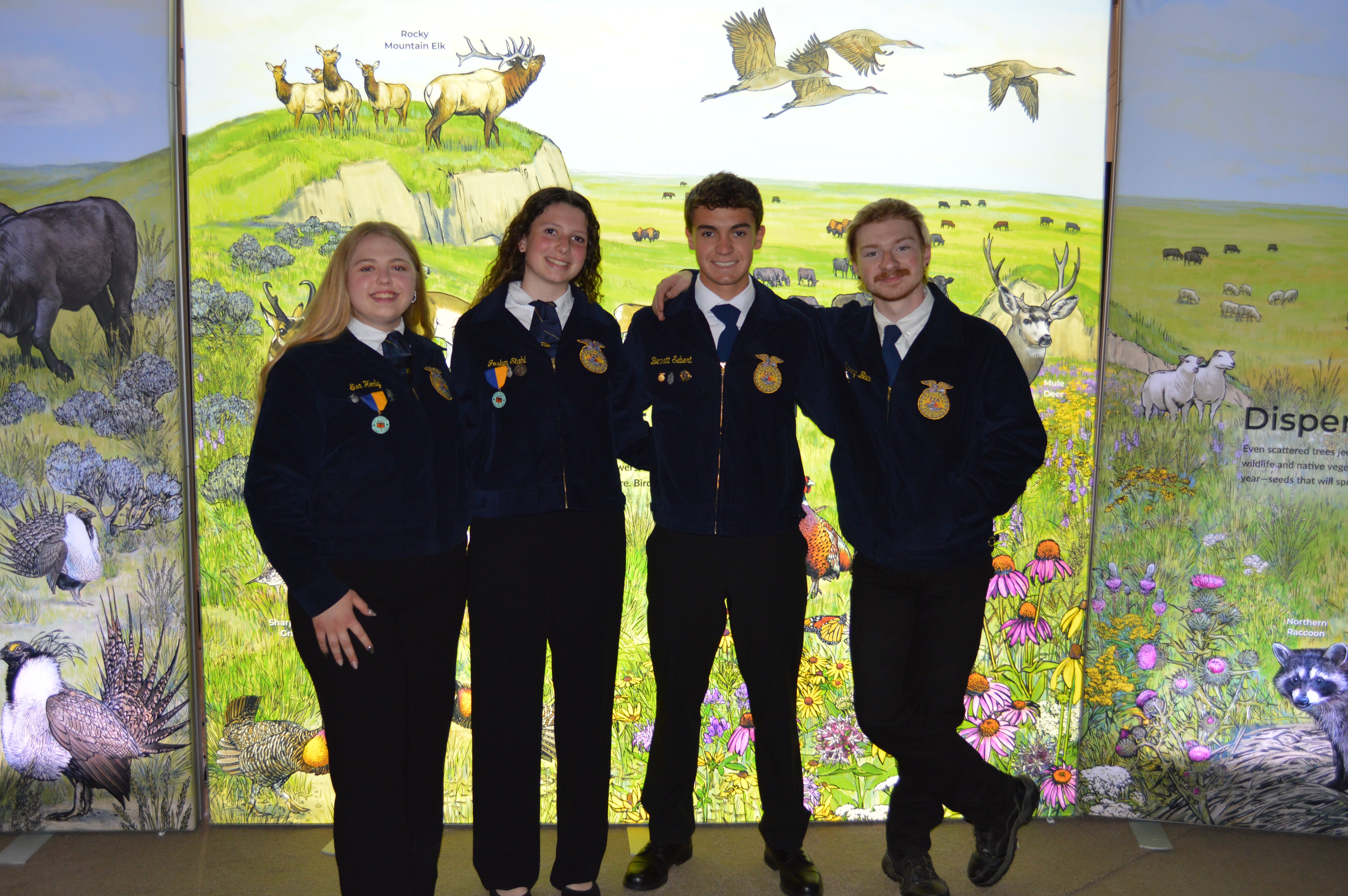 Four youth in matching FFA jackets stand in front of a nature/wildlife mural