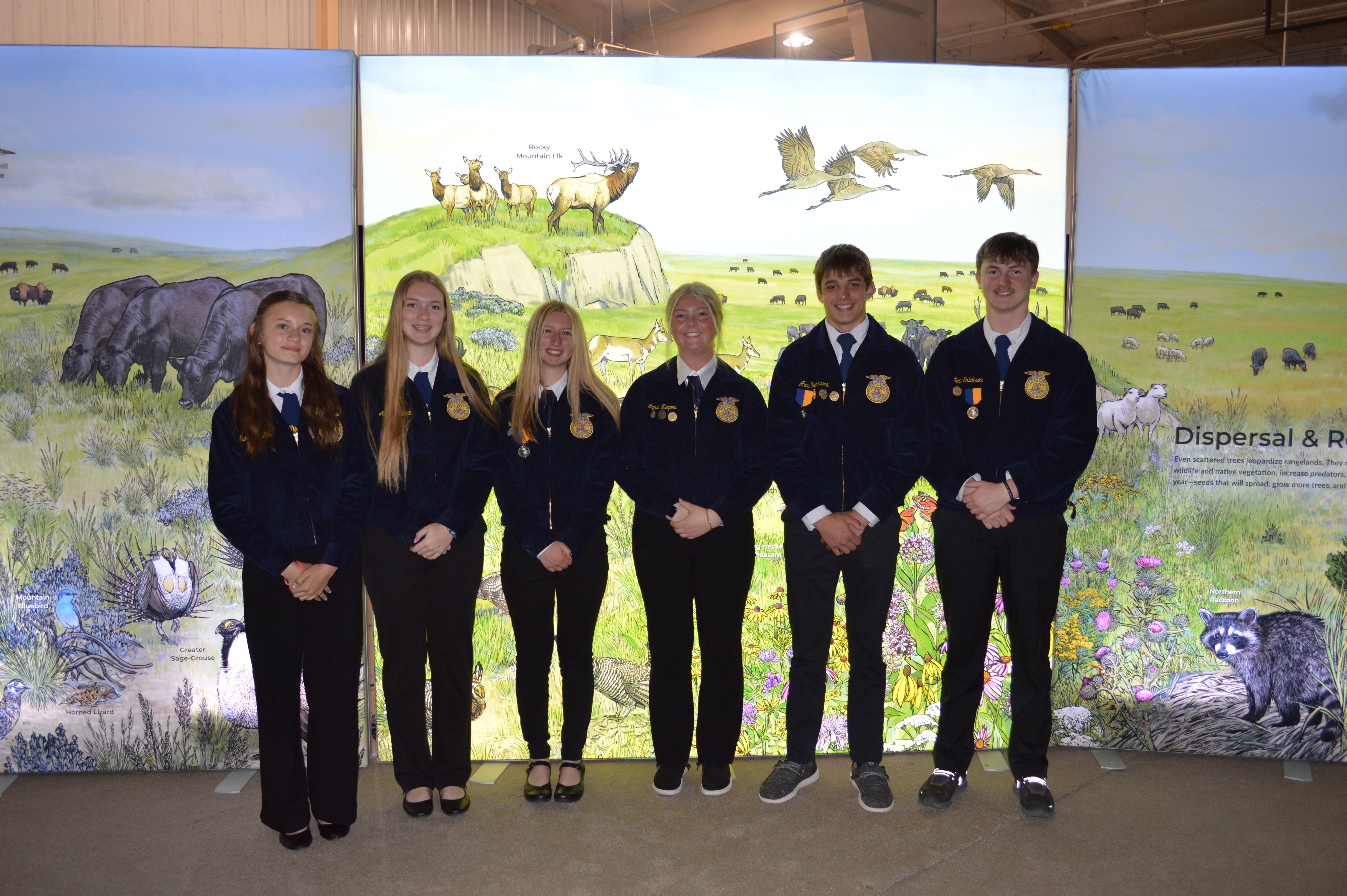 Six youth in matching FFA jackets smile in front of a nature/wildlife mural