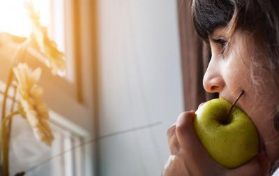 A young woman eating a green apple while watching the sun rise outside her window.