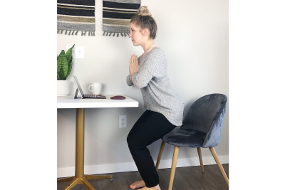 A young woman demonstrating the chair yoga pose. She is standing upright, slightly squatting with her hands folded in front of her.
