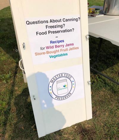 Sign outside Tim Shreiner's food preservation booth. The sign features the master food preserver logo, and reads: Questions about canning, freezing, food preservation? Recipes for Wild Berry Jams, Store-bought fruit jellies, vegetables.