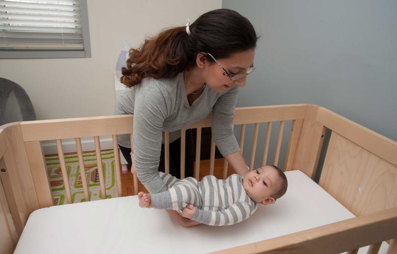 A young woman carefully placing her infant child in a crib on his back.