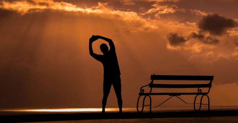person stretching in front of the rising sun near a bench on the beach