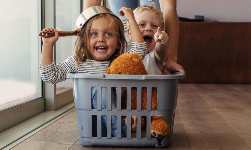 Mother pushing laundry basket along the floor with two children riding inside it.