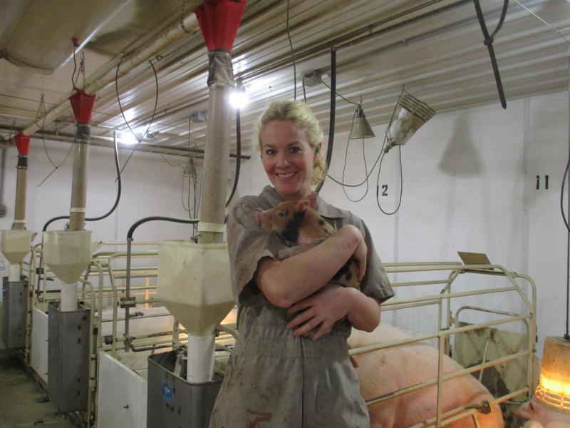 A woman standing inside a pig barn and holding a piglet