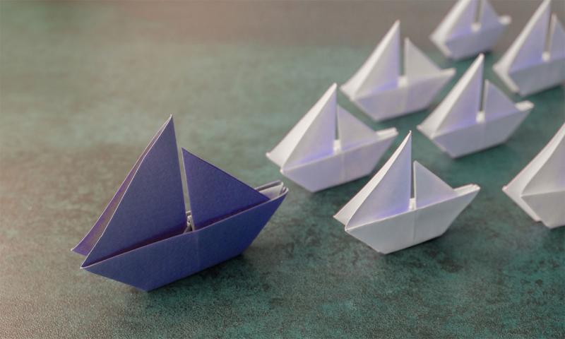 Purple origami paper sailboats leading several smaller, white paper sailboats.