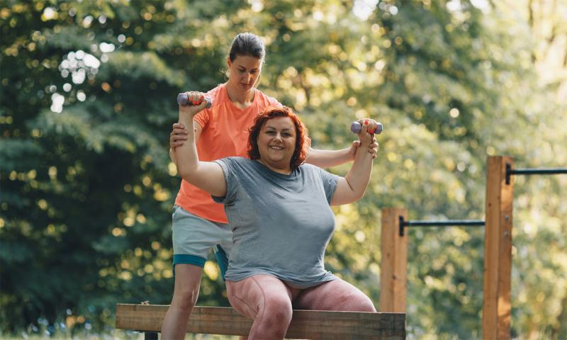 Female fitness instructor assisting an adult female at an outdoor gym.