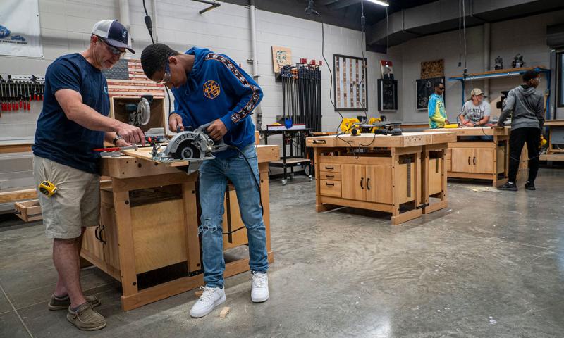 Man helping a younger man cut a piece of wood to size in a workshop.