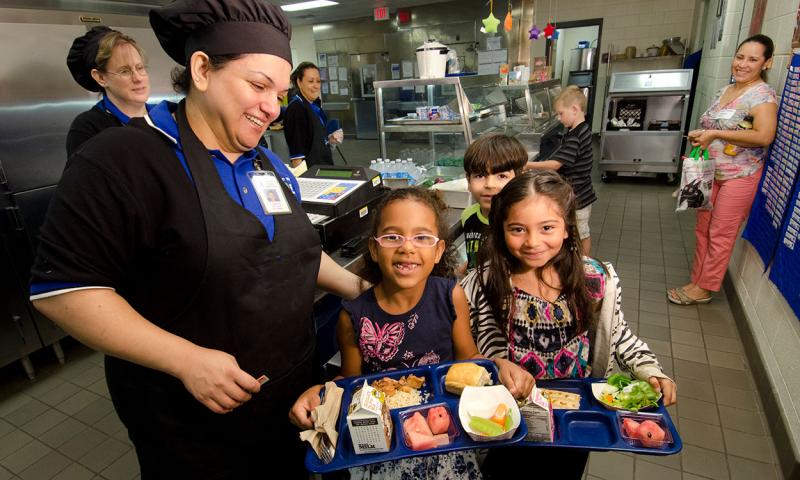 School lunch staff serving trays of healthy food a group of children.