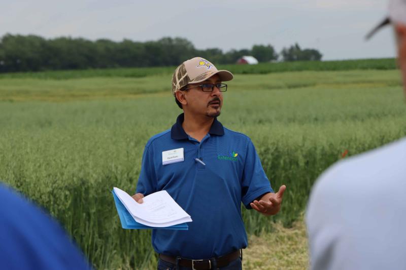 David Karki speaks to the audience at Northeast Field Day. He is standing outside wearing a blue shirt and a baseball cap.