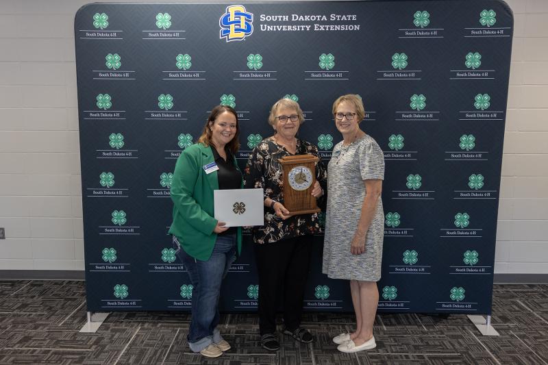 three women smile at the camera, with a 4-H backdrop behind them. The woman in the middle holds a commemorative plaque