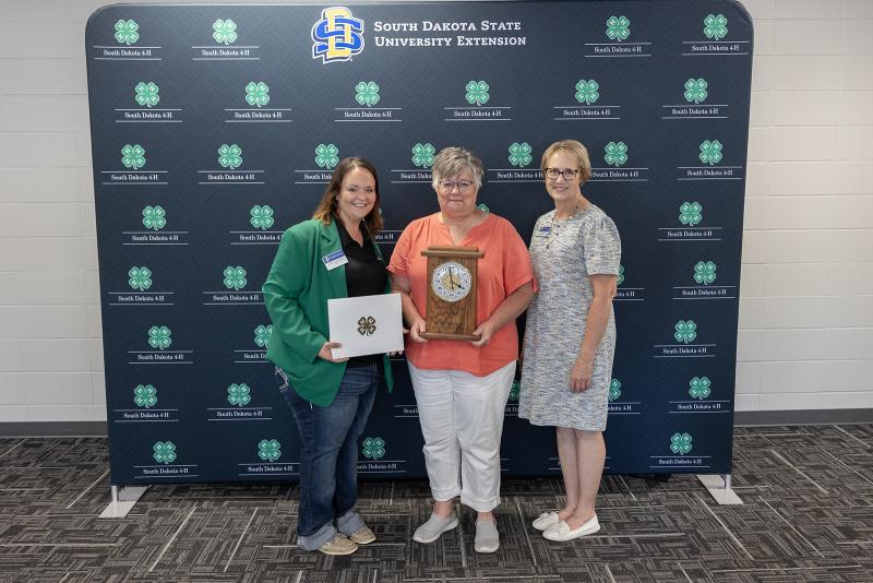 three women smile at the camera, with a 4-H backdrop behind them. The woman in the middle holds a commemorative plaque