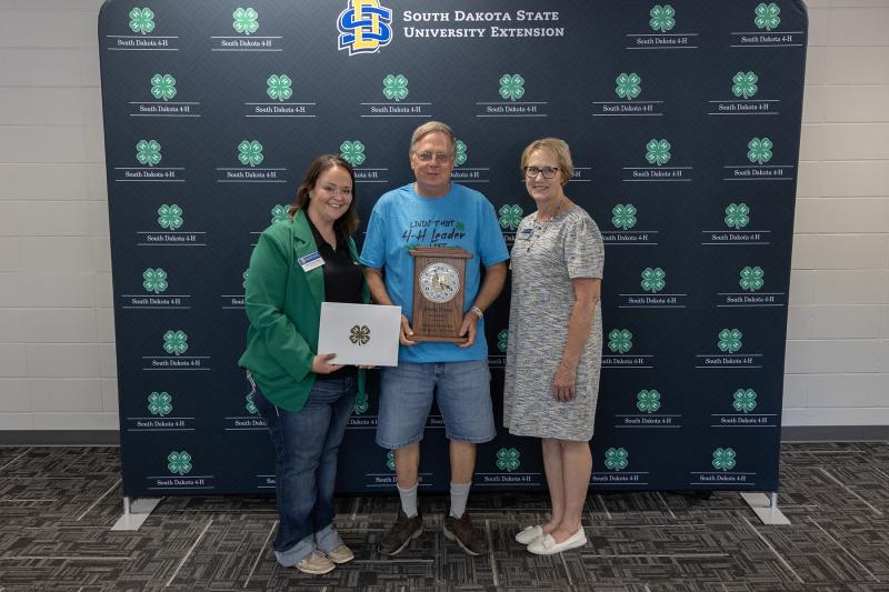 three people smile at the camera. the man in the middle holds a commemorative plaque