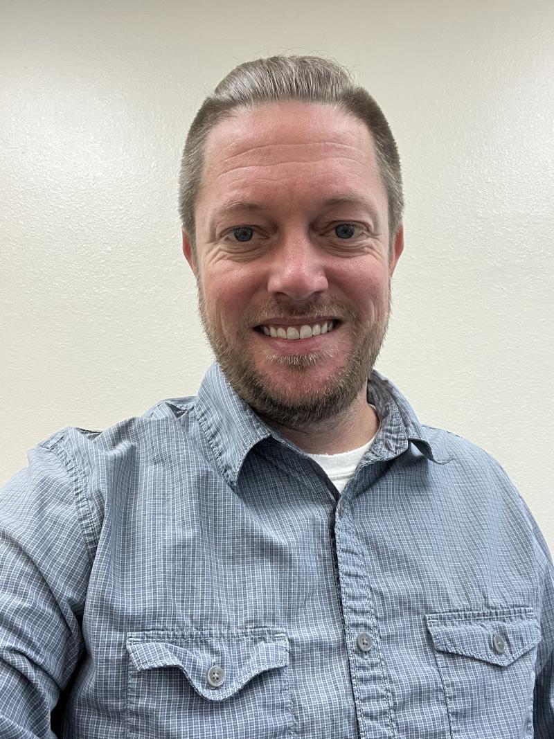 a man in a blue shirt with light brown hair and a light beard smiles at the camera
