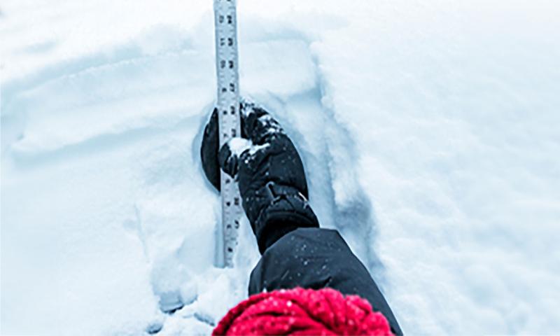 A woman estimating snow depth on a roof using a stiff ruler.
