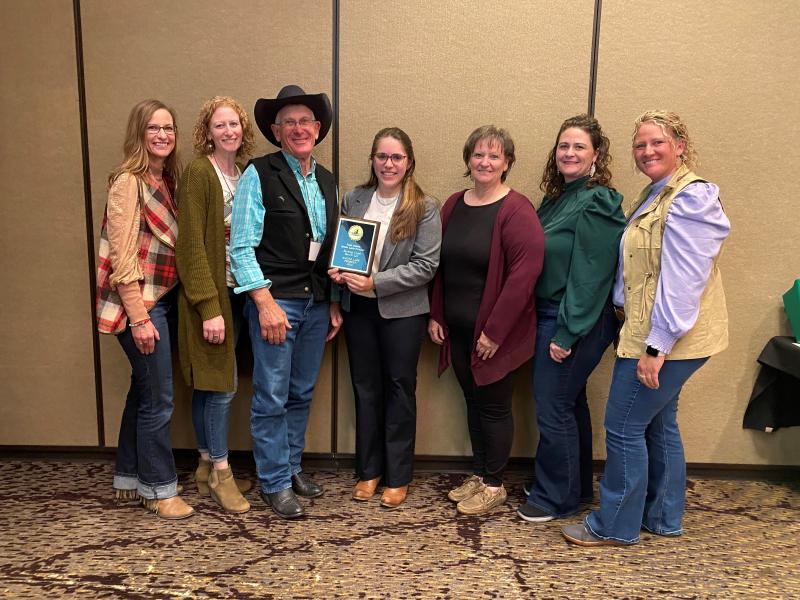 a woman with long brown hair presents a plaque to a man in a black cowboy hat. five women also pose and smile at the camera