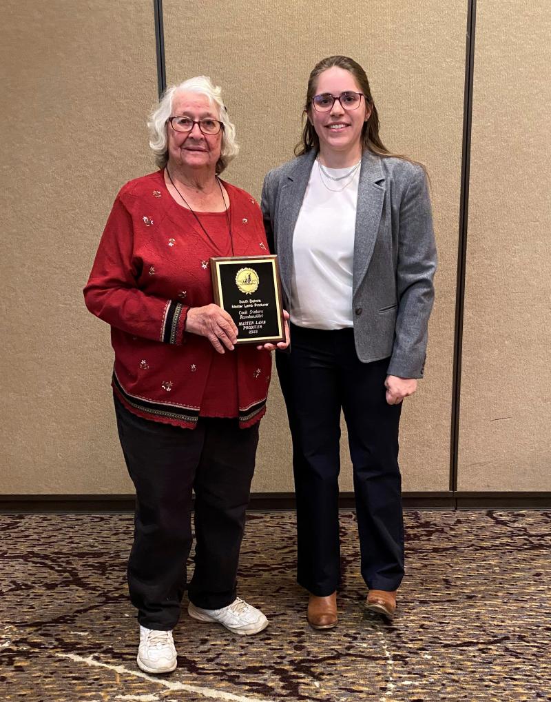 a woman with white hair and glasses holds a plaque for the 2023 Master Lamb Producers Award next to a woman with long brown hair wearing a suit jacket and dark slacks