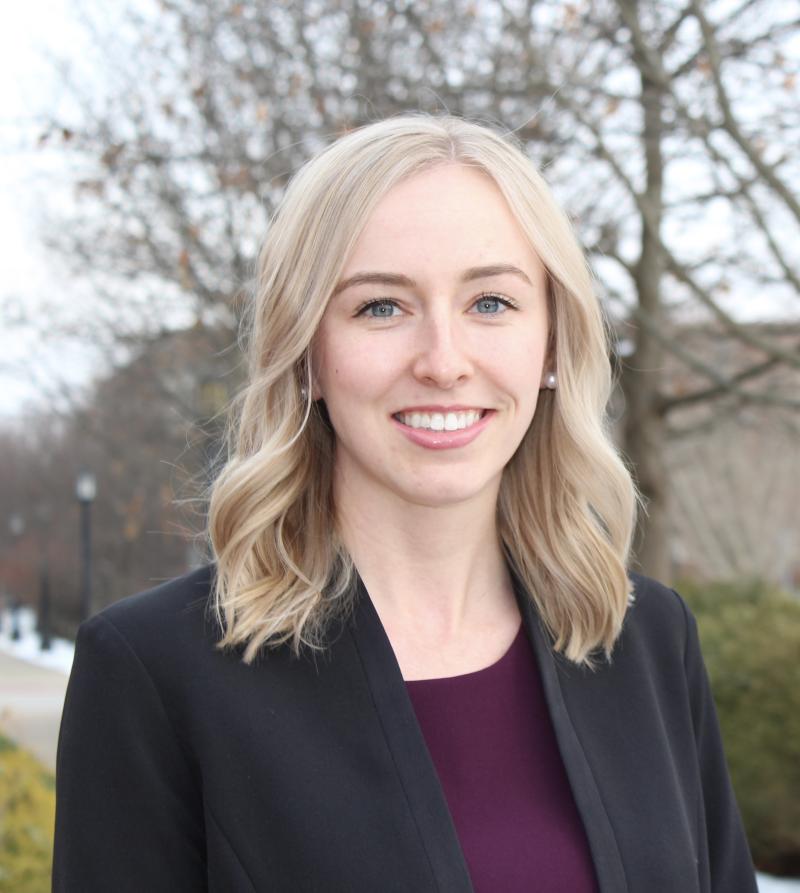 Sarah Sellars smiles at the camera. Her shoulder-length blonde hair is down, and she is wearing a purple top with a black blazer
