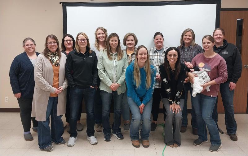 The Britton graduating class of Annie’s Project and event organizers stand for a group photo in front of a white screen. There are 13 women, plus one woman on the far right holding a baby
