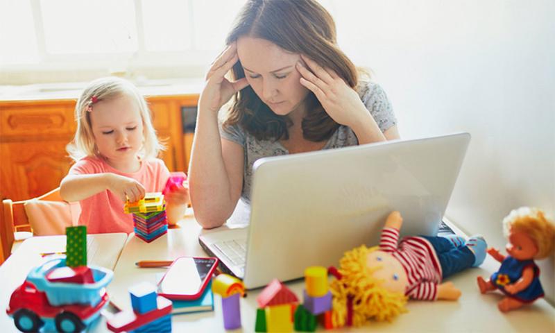 Tired mother working on her laptop with a toddler at her side.