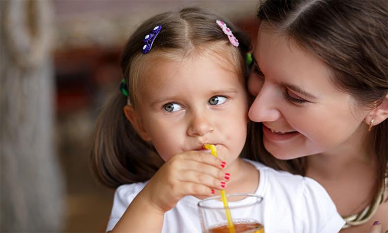 Young mother smiles at her toddler daughter as she sips on a glass of juice.
