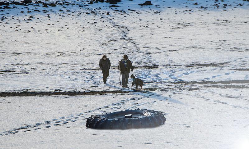 Two ranchers with a dog inspecting a cattle tire waterer in a snow-covered pasture.