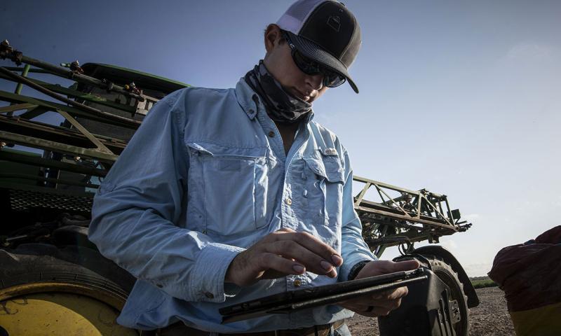 Young farmer analyzing data on a tablet computer beside a crop sprayer.