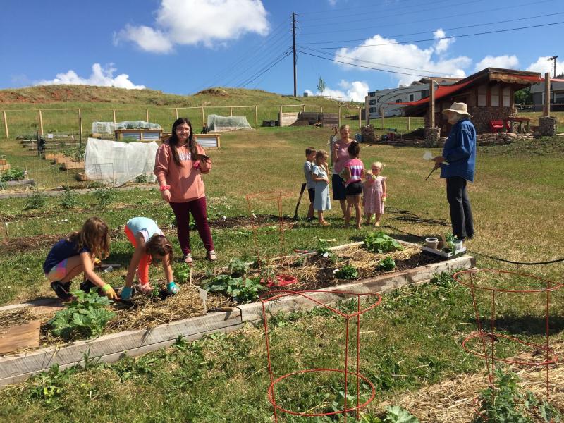 Two children work in a garden bed while off to the right a women speaks to a small group of children
